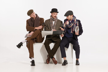 Portrait of three stylish men, gentlemen sitting together and looking on phone in magazine isolated over grey background