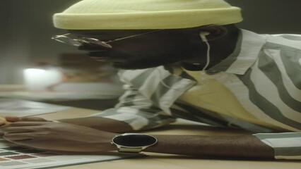 Vertical close up shot of African American male designer listening to music, using color palette and drawing picture in copybook while working on creative project at night - Powered by Adobe