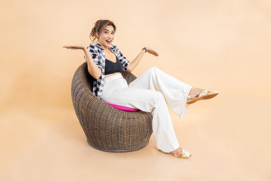 Happy Indian Woman Wearing Casual Cloths Looking At Camera While Sitting In Chair With Surprised Expression And With Open Arms Gesture Isolated Over Beige Background.