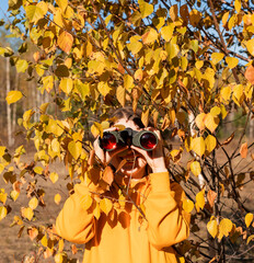 happy Young woman in yellow hoodie with binoculars watching birds in the autumn forest...