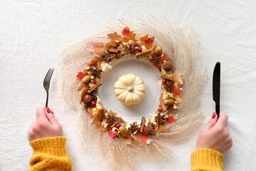 Autumn wreath and white pumpkin on a plate. Hands with kitchen utensils, fork and knife. Fall flat...