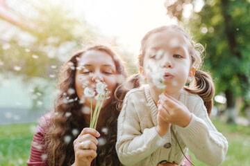 Young pretty women with small girl blowing dandelions in the park. Spend happy time together outside.