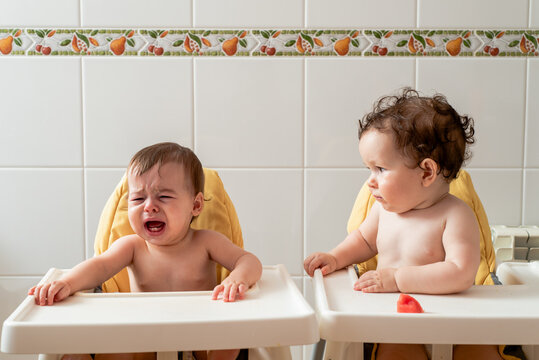 Cute baby crying while sitting in dining chair near twin sibling