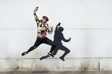 Black man jumping and taking selfie near wall