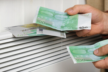 Human Hand Holding Euro Banknotes near Electric Heater Radiator at Home. Man Pay Money for Heating. Expensive to Heat. Energy Saving. Rising Cost of Living. Global Gas Crisis. Paying Cold Season Bills