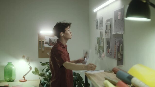 Gen Z Asian interior designer pinning printed pictures to wall while creating mood board at office workplace