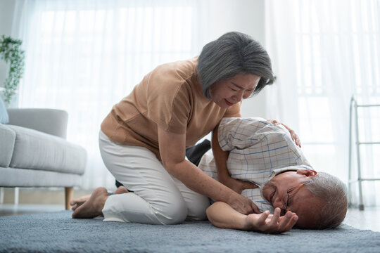 Asian Senior Wife Helping Husband Fainting And Falling On The Ground In The Living Room. Elderly Male Patient Having An Accident Or Heart Attack. Home Nursing And Health Insurance Concept