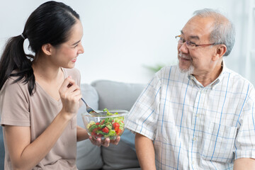 Happy Asian daughter making salad and feeding to senior father sitting on couch at home. Senior man smiling of tasty food from beloved girl. Family spending time together on weekend