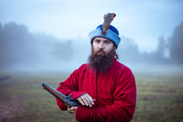 Bearded man in 17th century clothes and hat with antique pistol cocks the trigger