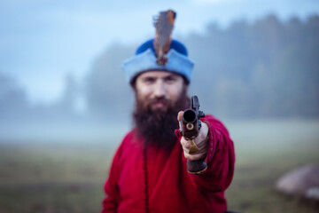 Bearded man in 17th century clothes and hat with antique pistol aims directly at the camera, selective focus on the muzzle of the gun