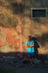 Blond man in blue shirt and swim shorts stands against weathered pink plaster wall in late afternoon sunlight.