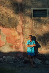 Blond man in blue shirt and swim shorts stands against weathered pink plaster wall in late afternoon sunlight.