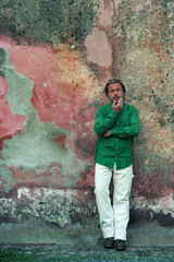 Obraz premium Blond tanned man in green shirt and white pants stands in front of weathered pink plaster wall.