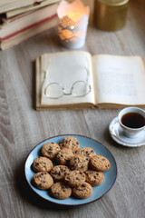 Plate of chocolate chip cookies, stack of vintage books, reading glasses, cup of tea or coffee, lit candle and fairy lights. Hygge at home. Selective focus.
