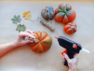 Women's hands with a hot gun stick a twine tail to artificial alabaster pumpkins on a beige background