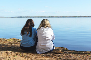 Outdoors, two women dressed in a blue hoodie and a white jacket with flowing hair are sitting on the shore of a reservoir, looking from the back and looking ahead at the horizon, copy space.