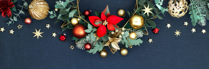 Decorative Christmas border. Panorama with floral garland with eucalyptus, baubles, trinkets and red poinsettia. Red, green and golden Xmas decorations on dark blue linen background, panoramic banner.