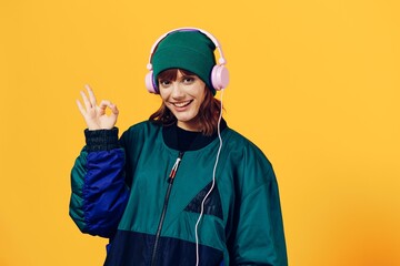 a joyful woman in a green jacket and a hat in the style of the 90s enjoys music standing in pink headphones on a yellow background showing the ok sign while looking at the camera