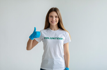 I like volunteering. Positive lady in white volunteer t-shirt showing thumb up, wearing blue gloves, light background