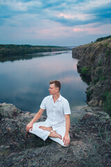 Fototapeta premium A handsome young guy is engaged in meditation sitting in a lotus position at dawn, morning meditation on the river bank.