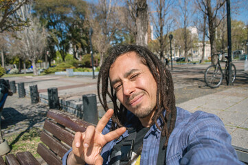 young latin man with dreadlocks in a square takes a selfie smiling and making a v sign with his hand