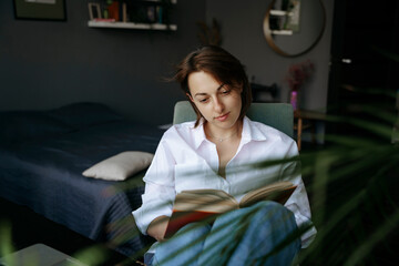 A woman is sitting at a desk in a home office and reading a book. In the background is her bedroom, and next to it is a laptop