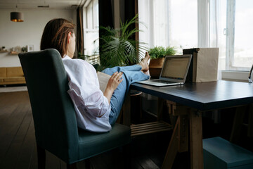 A woman is sitting at a desk in a home office and reading a book, there is a laptop on the table. 