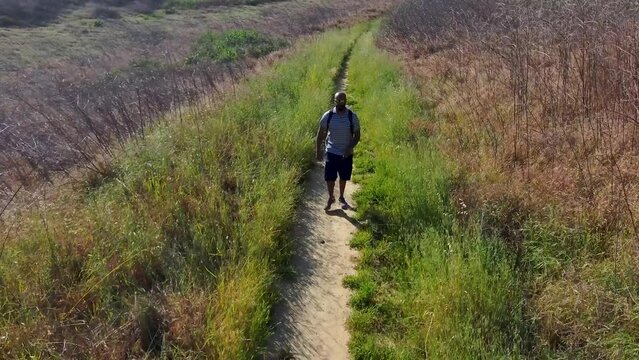 aerial footage of an African American man walking along a footpath in Malibu Creek State Park surrounded by majestic mountain ranges covered in dry brush and lush green plants in Chino Hills