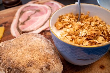 A bowl of muesli and yogurt and a roll of bread with ham.