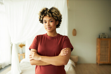 Horizontal indoor portrait of african american woman standing in bedroom with confident smile looking at camera with folded arms, wearing red t-shirt. Happy real estate owner. Daily life