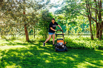 Brown-haired female gardener in summer casual clothes using gasoline lawn mower or grass cutter in the yard, grounds. Technological machine tool for lawns. Woman caring her garden against flower trees