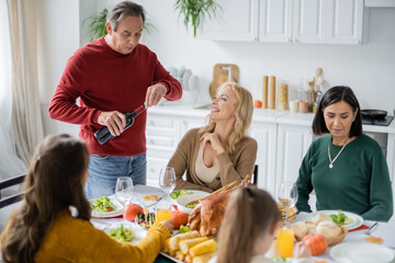 Smiling woman looking at parent with wine near thanksgiving dinner and multiethnic family at home