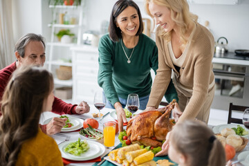 Multiethnic mother looking at daughter holding turkey near blurred family and thanksgiving dinner at home