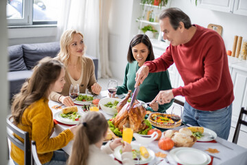 Smiling woman looking at parent cutting thanksgiving turkey near multiethnic family at home