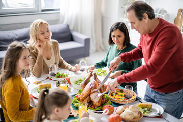 Smiling multiethnic family looking at senior man cutting thanksgiving turkey at home