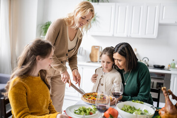 Smiling woman cutting thanksgiving pie near multiethnic mother and daughter at home