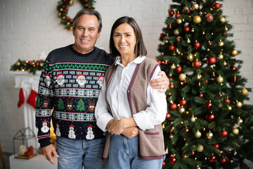 smiling man in sweater with festive pattern hugging multiracial wife near blurred christmas tree