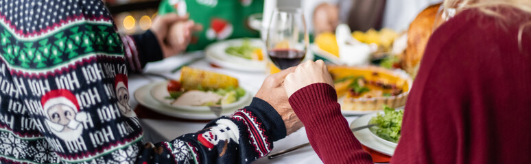 partial view of senior father and adult daughter holding hands and praying near blurred thanksgiving dinner, banner