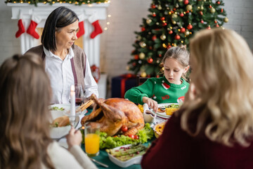 multiracial woman looking at granddaughter during festive dinner with blurred family near decorated christmas tree