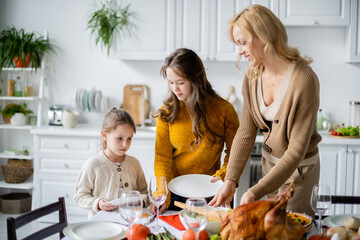 smiling blonde woman serving table for thanksgiving dinner with daughters in kitchen