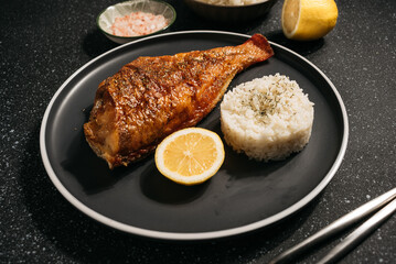baked fish perch with lemon, boiled rice, spices nearby. fish on a black plate and dark background
