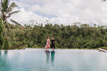Fototapeta na wymiar Brunette woman in pink dress holding hands with friend during photoshoot at Bali. Outdoor photo of female models standing near pool on jungle background.