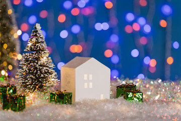 Christmas cute little house with tree in snow at night. Glowing festive garland on violet background with bokeh lights. Cozy atmosphere with home decor with selective focus. New Year card