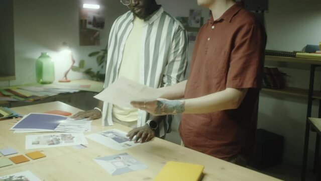 Asian and African American designers discussing printed pictures while working together on creative project during night in studio
