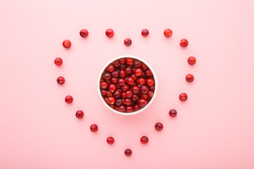 Heart shape created from red berries. Fresh cranberries in white bowl on light pink table background. Pastel color. Love healthy food. Closeup. Top down view.