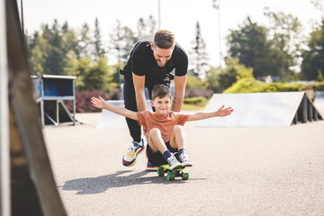 young boy learning to ride skateboard as father teaches him in the suburb street having fun.