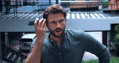 Handsome curious pensive multi-ethnic dark-haired young man, in casual wear, standing outdoor and holding his mobile phone at ear level and experiencing shock while listens to a recorded voice message