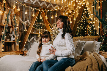 Fototapeta na wymiar A little girl with her mother in a cozy home environment on the sofa next to the Christmas tree. The theme of New Year holidays and festive interior with garlands and light bulbs
