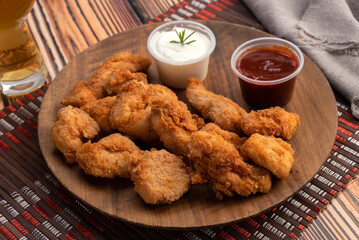 Chicken nuggets on a round wooden plate with two bowls of red sauce and white cheese sauce and glass of beer