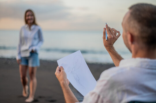 Young couple on a beach at sunset, she poses and he draws a picture of her